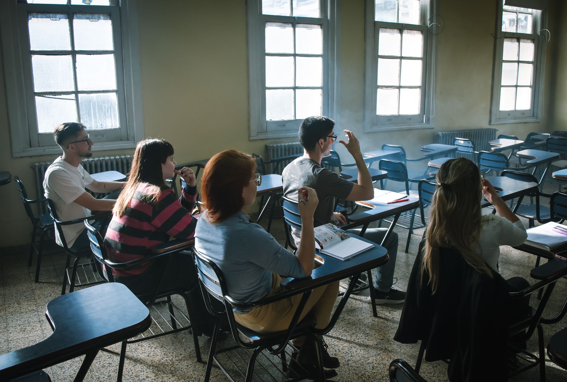 College students at desk chairs in a sunlit classroom, one raising a hand during a lecture