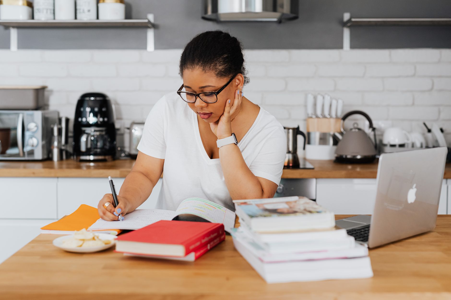 Woman taking notes at a kitchen counter with a stack of textbooks and a laptop beside her