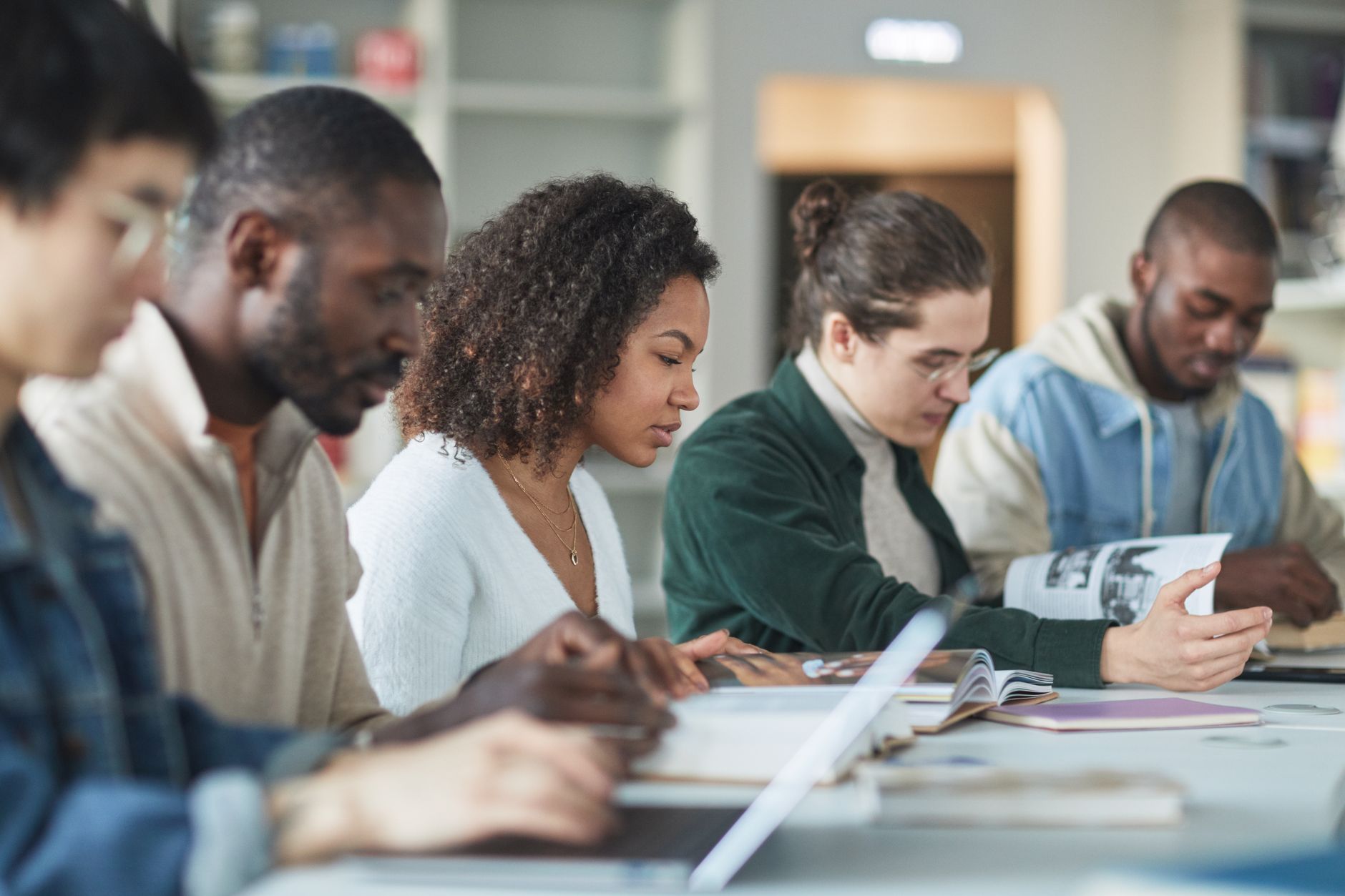 A row of college students reading textbooks and taking notes on laptops at a long library table
