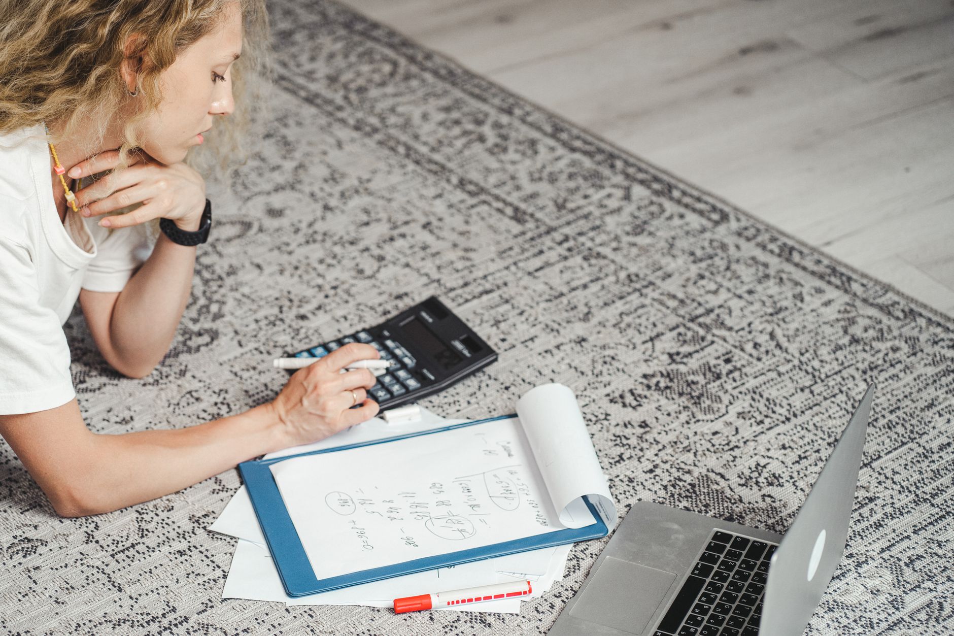 A woman lying on a rug working through figures with a calculator, a clipboard of notes and an open laptop