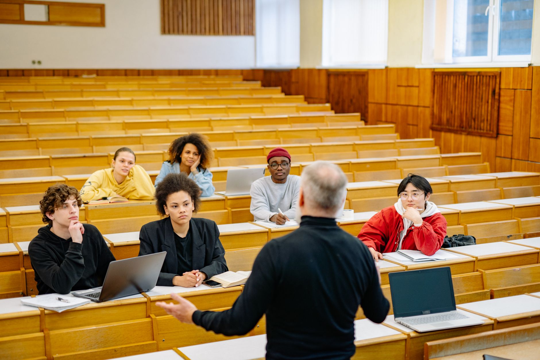 A professor in a black sweater speaking to a small group of students seated with laptops and notebooks in a lecture hall