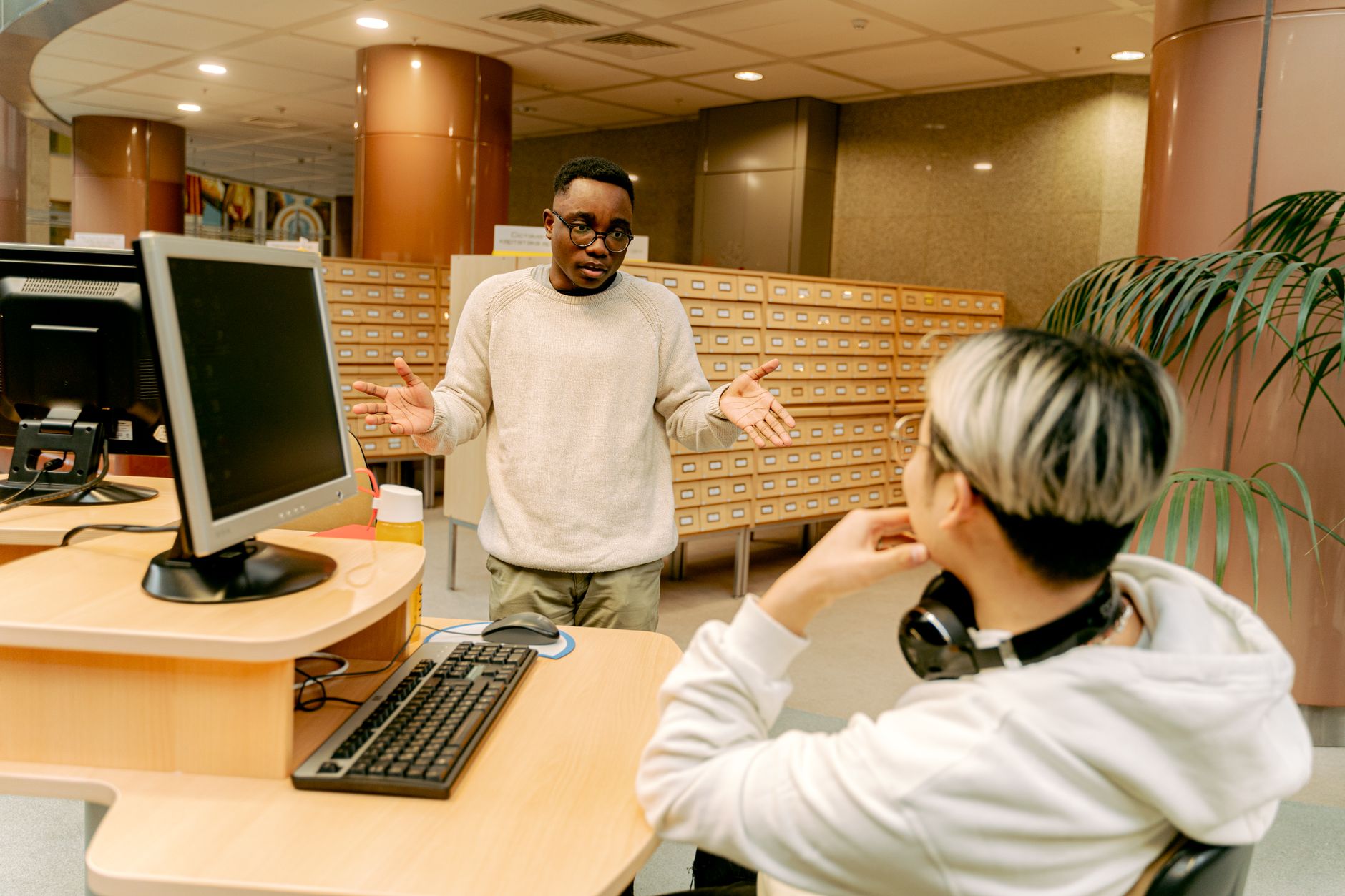 Two college students talking at a computer desk in a university library, one standing and one seated