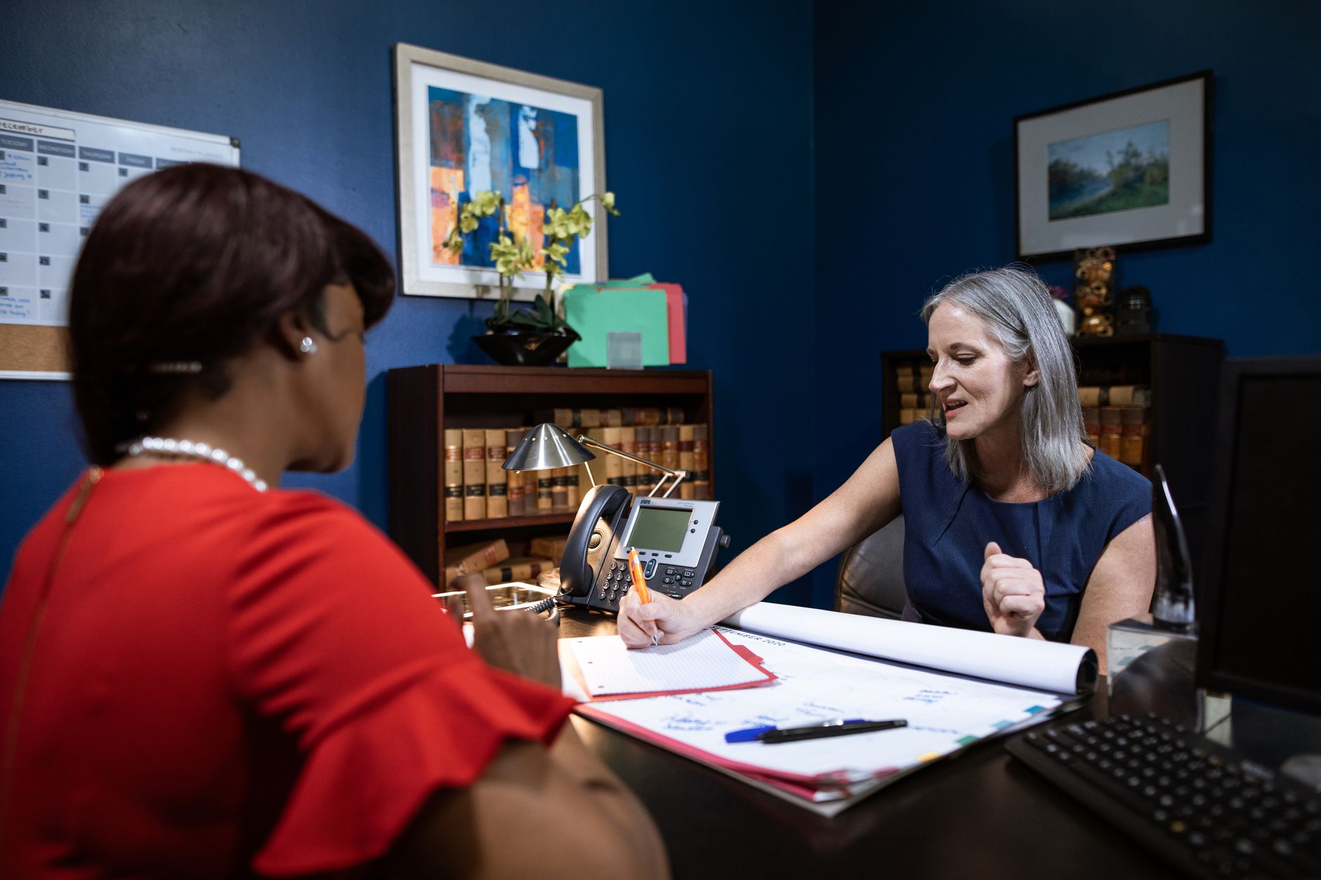 A case manager writes notes at her desk while meeting with a client across from her in a blue office