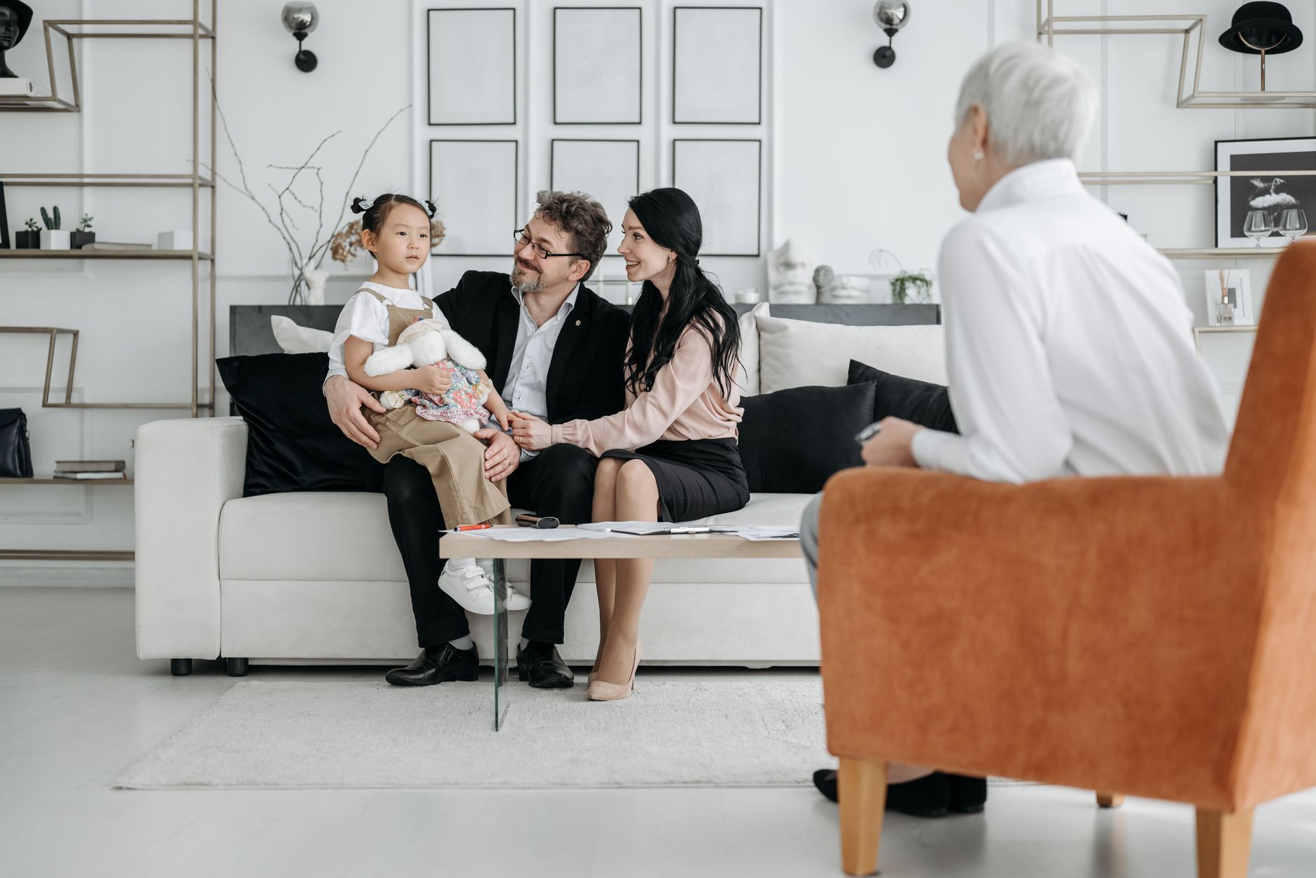 A social worker in an armchair meets with two parents and their young daughter on a living room sofa, with forms on the coffee table