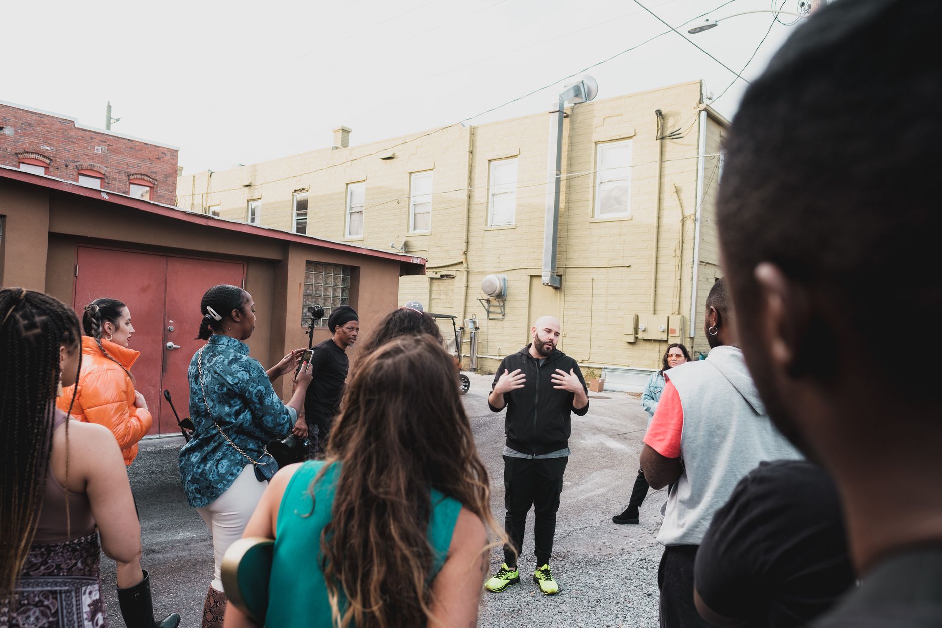 A community organizer speaks to a group of neighbors gathered in a back lot between brick and painted buildings