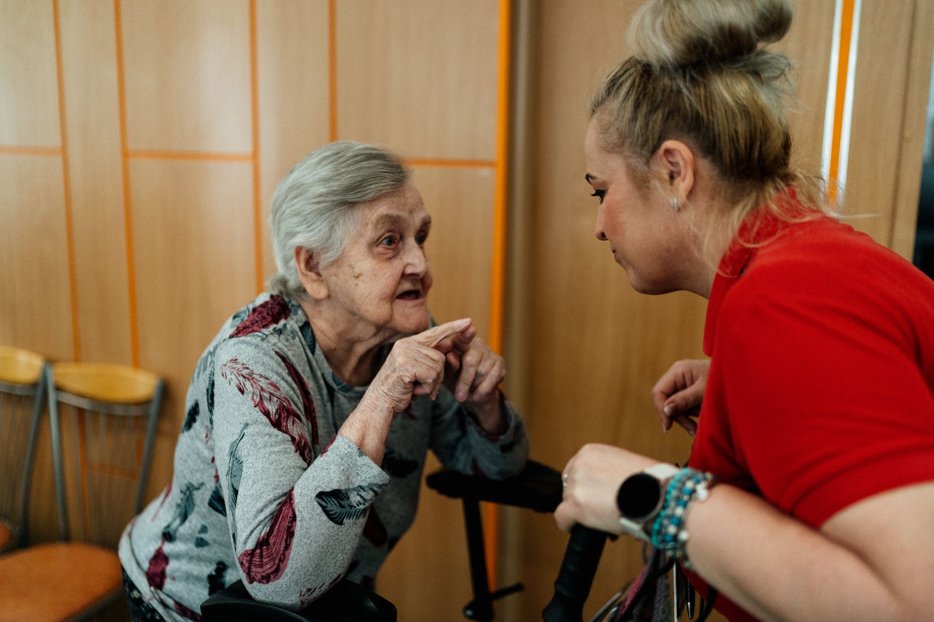 A care worker in a red shirt leans in to listen as an older woman using a walker talks with her