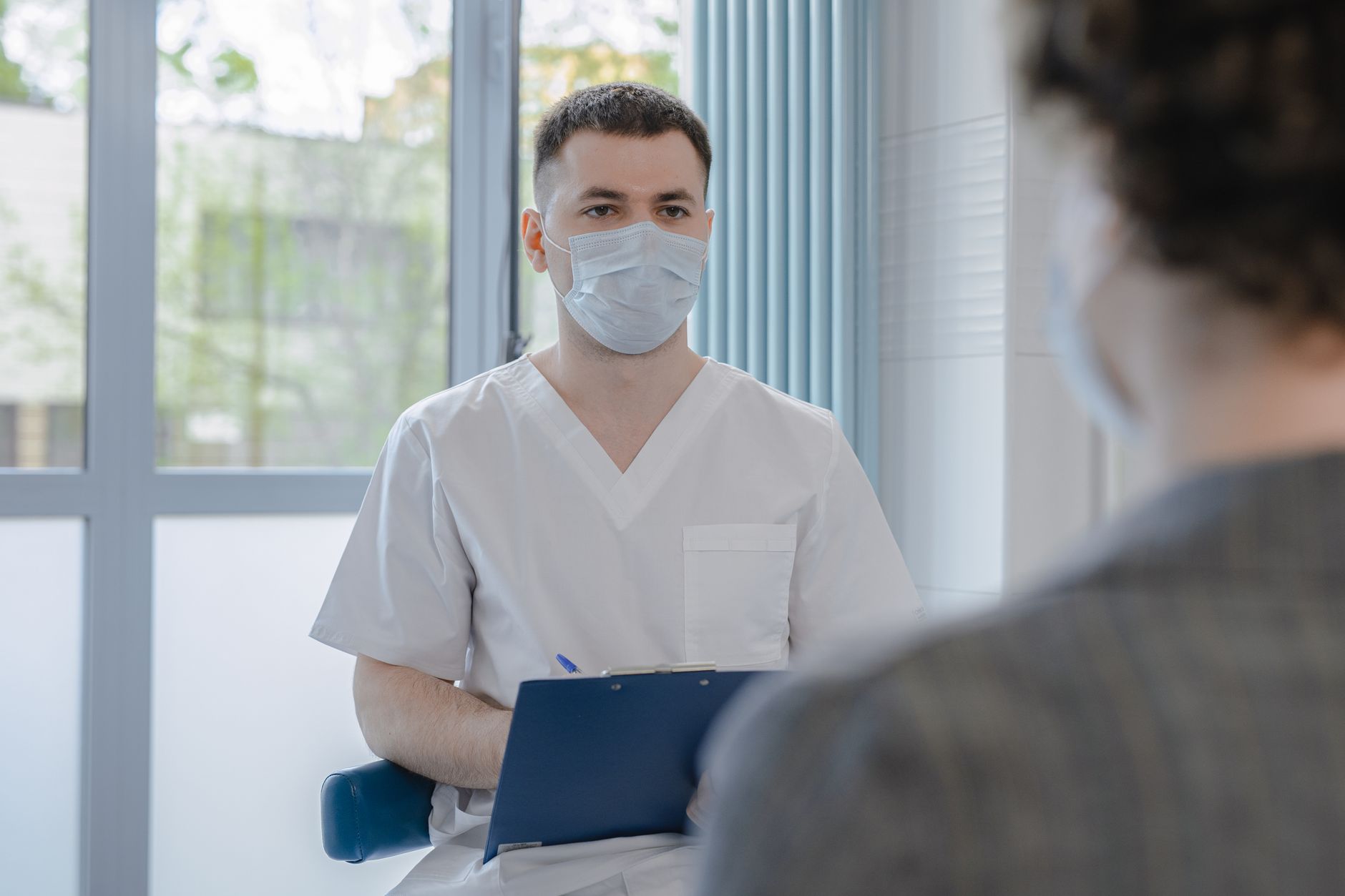 A health-care worker in scrubs and a face mask holds a clipboard while listening to a patient in a bright clinic room