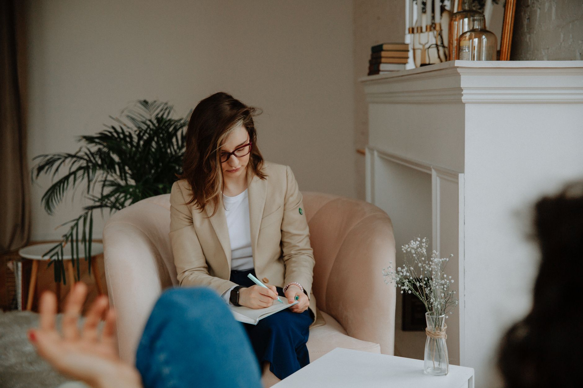 A counselor in a beige blazer writes notes in an armchair while a client, out of focus in the foreground, talks