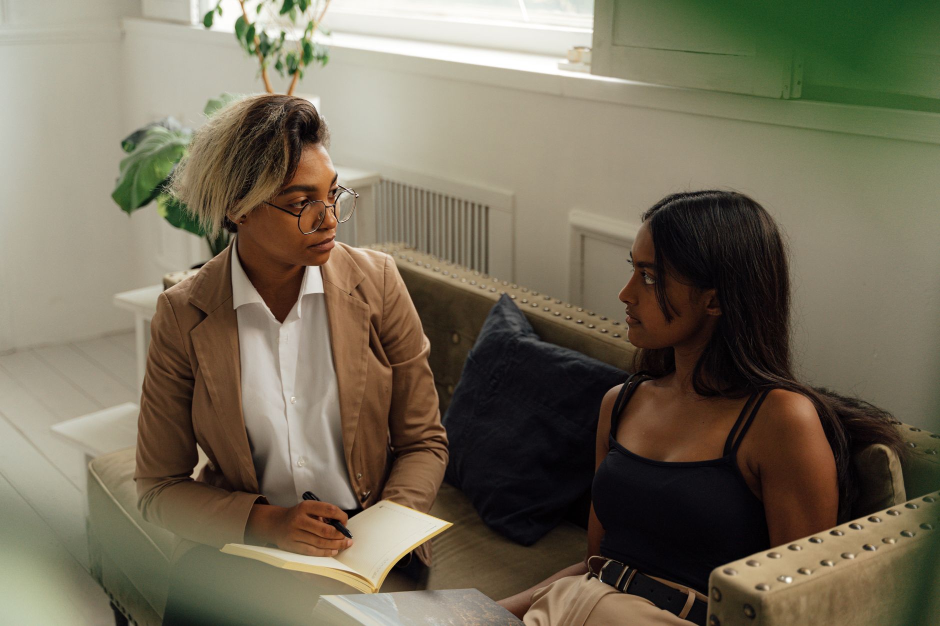 A counselor in a tan blazer takes notes in a notebook while talking with a young woman seated beside her on a sofa