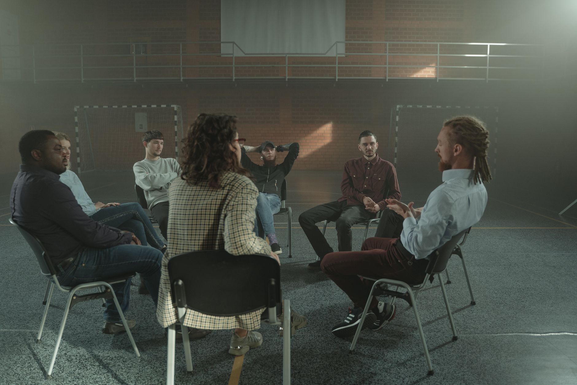 Eight adults sit on chairs in a circle in a gymnasium while a group leader speaks during a support session