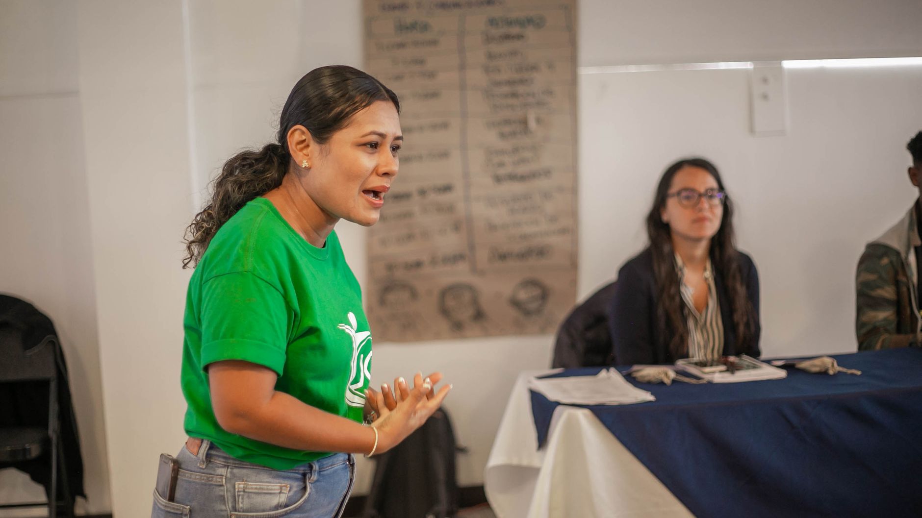 A woman in a green T-shirt speaking at a community workshop while a panel listens at a table behind her