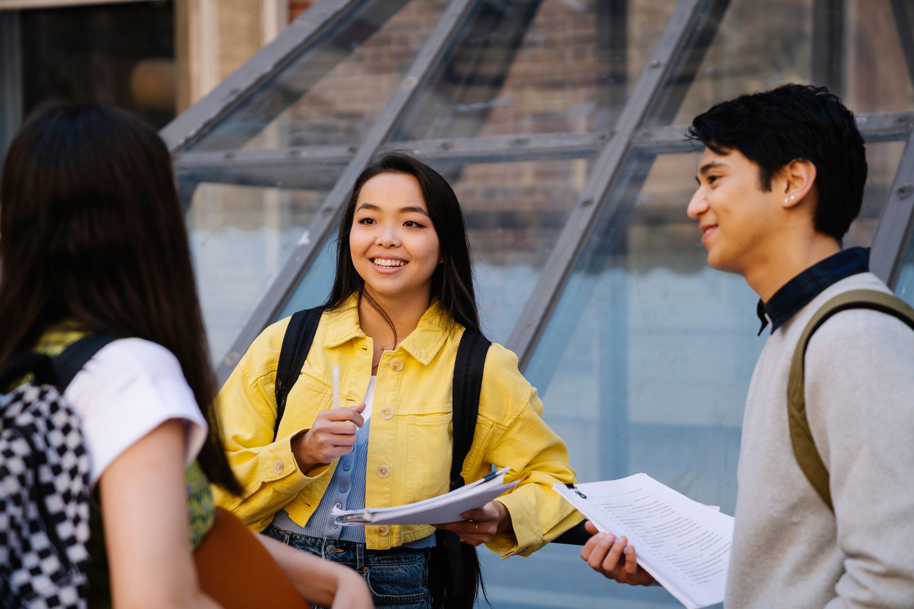 Three college students with backpacks talking outside a campus building, holding course papers