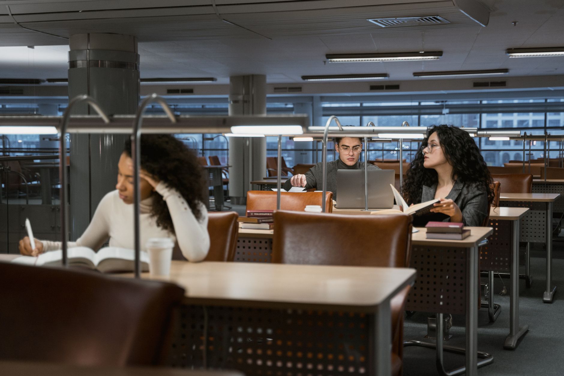 College students studying at reading-lamp desks in a university library, one taking notes from a textbook