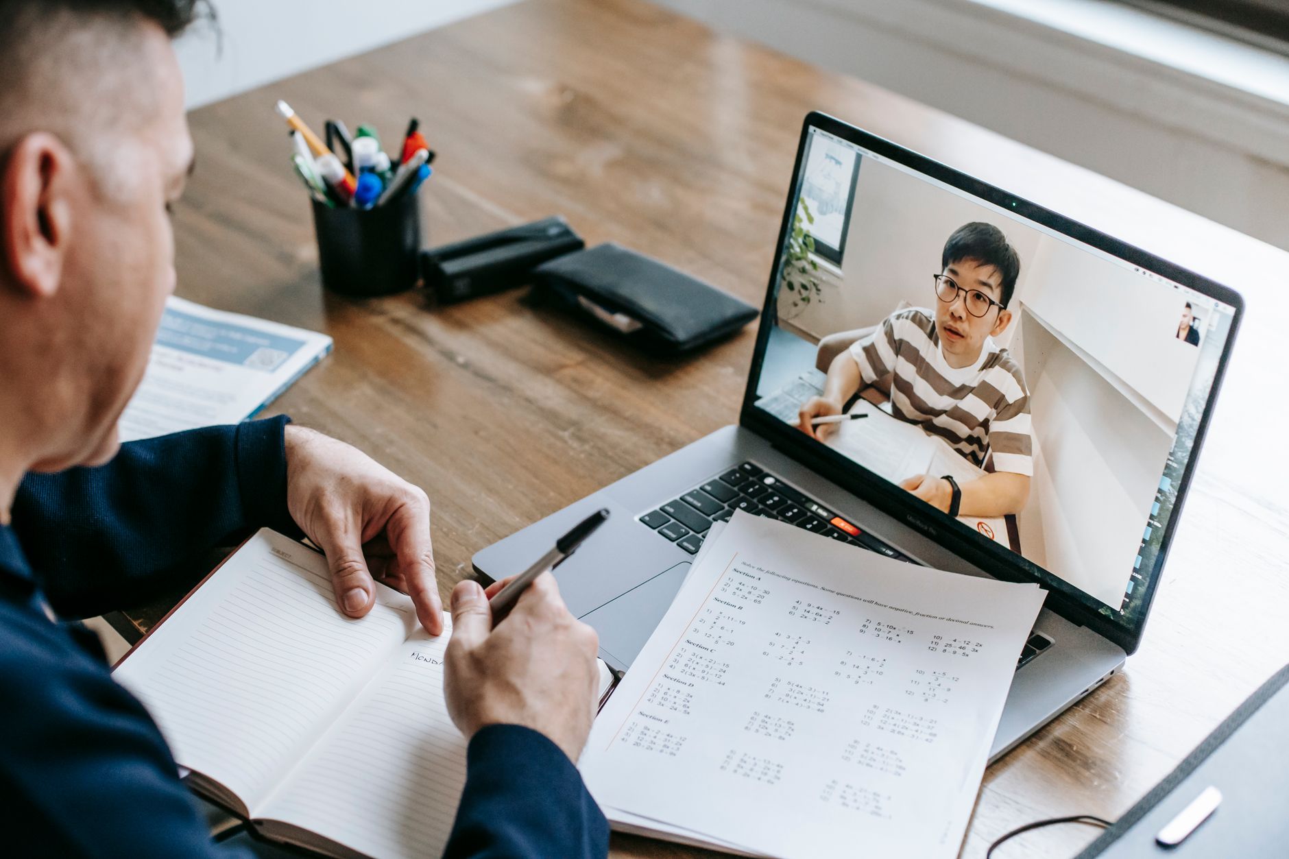 An adult student takes notes in a notebook during a video call shown on a laptop at a wooden desk